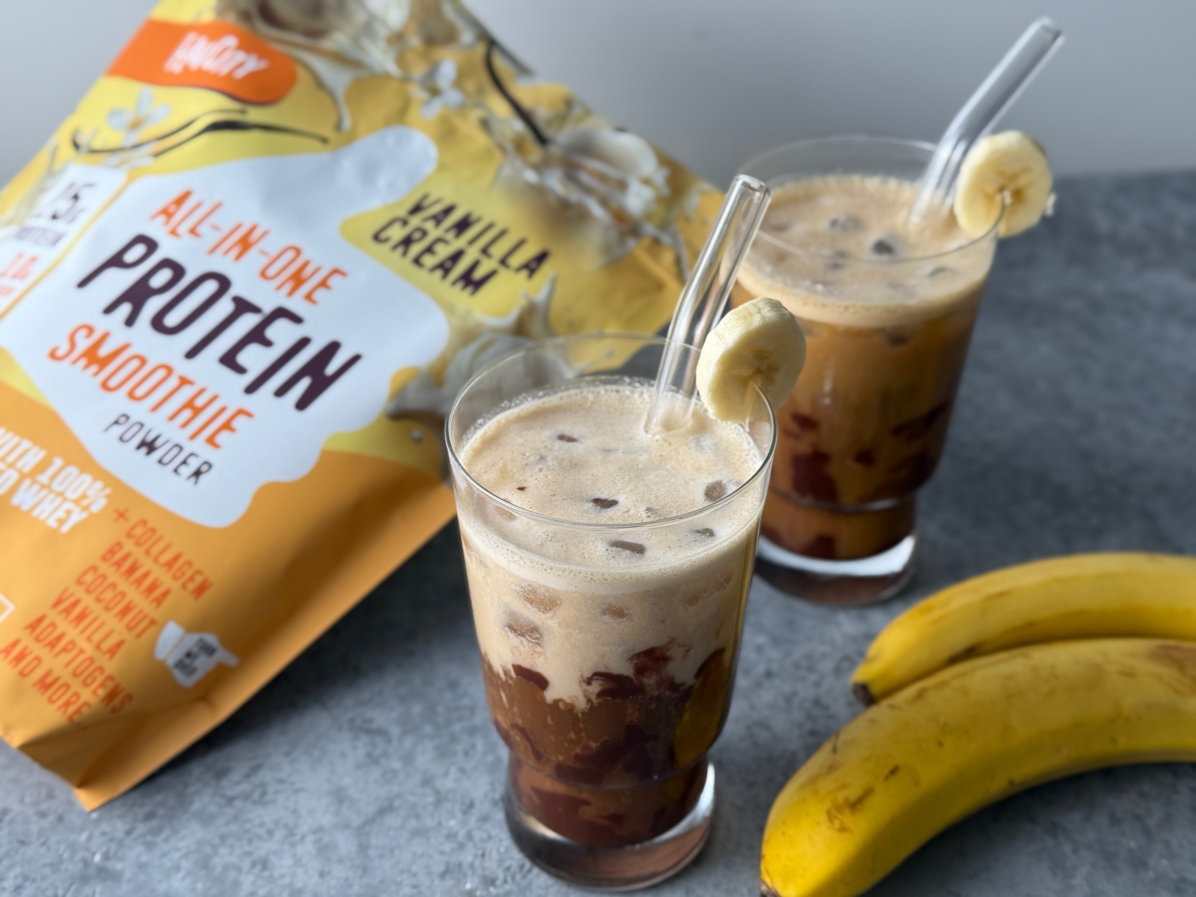 Two glasses of iced protein smoothies with glass straws and banana slices on the rim, placed next to a yellow bag of vanilla cream all-in-one protein smoothie powder and two ripe bananas on a gray surface